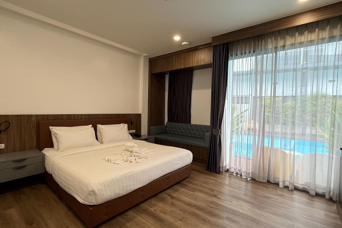 A hotel room with a large bed, a bedside table, a small desk, a sofa, and a view of a swimming pool.