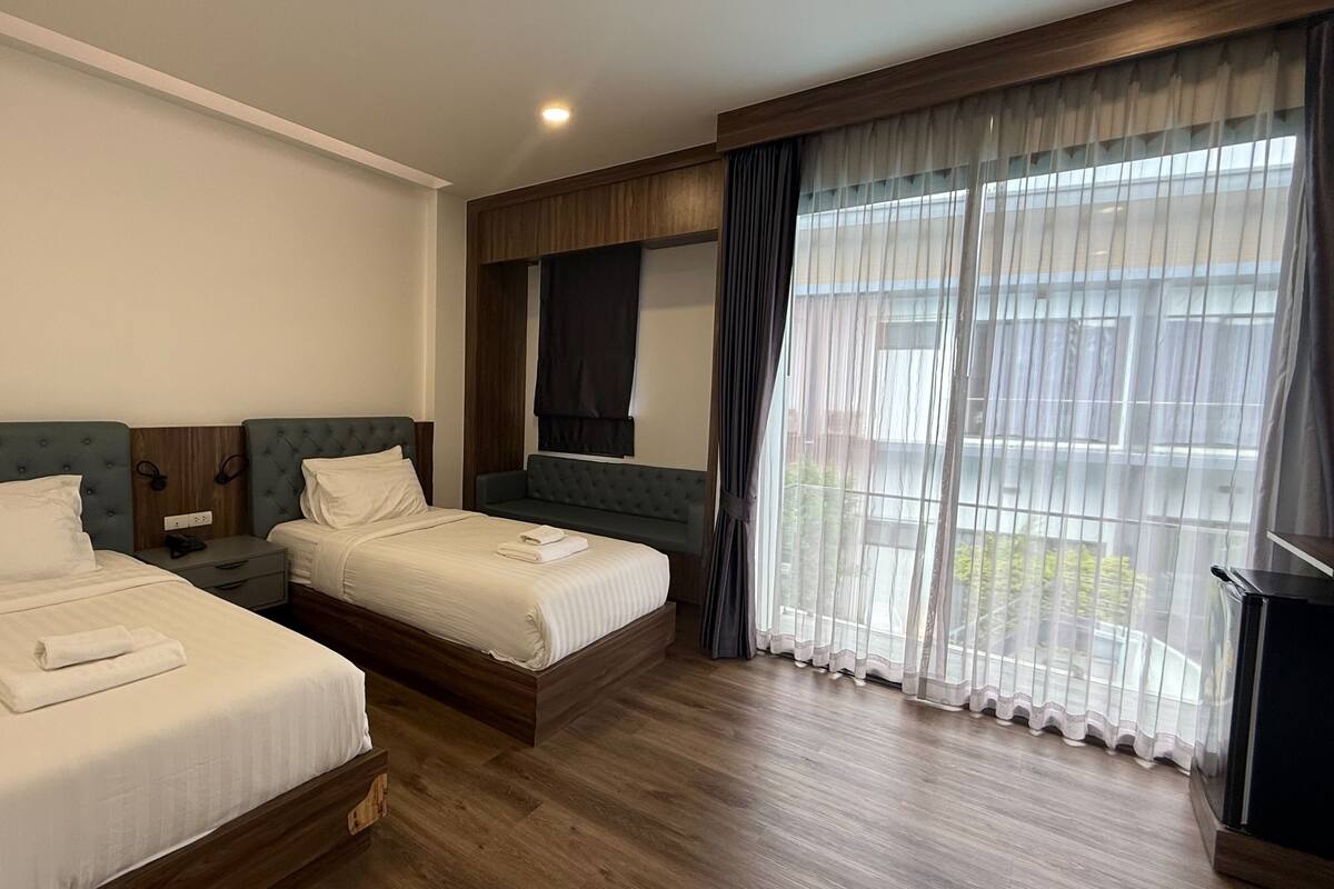 A hotel room with two beds, a wooden floor, a large window with curtains, and a mirror above the bed.