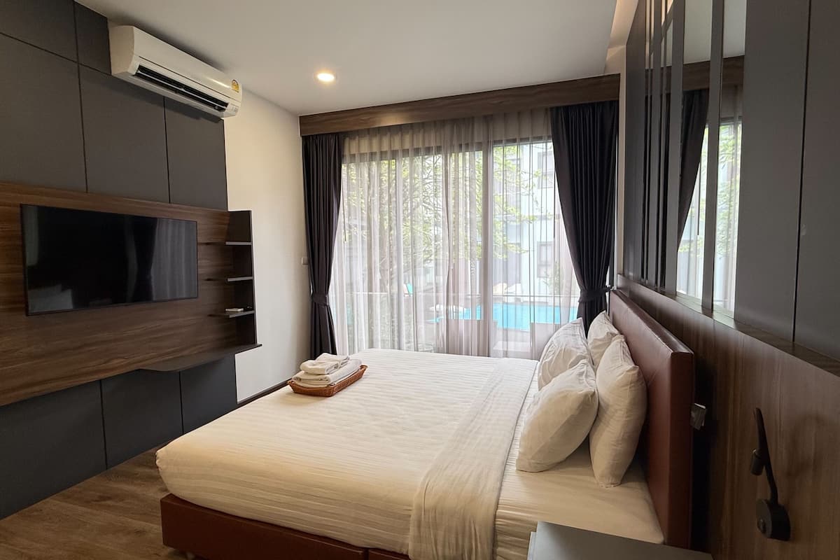 A modern hotel room with a bed, a flat-screen TV, a built-in air conditioner, and a view of a swimming pool.