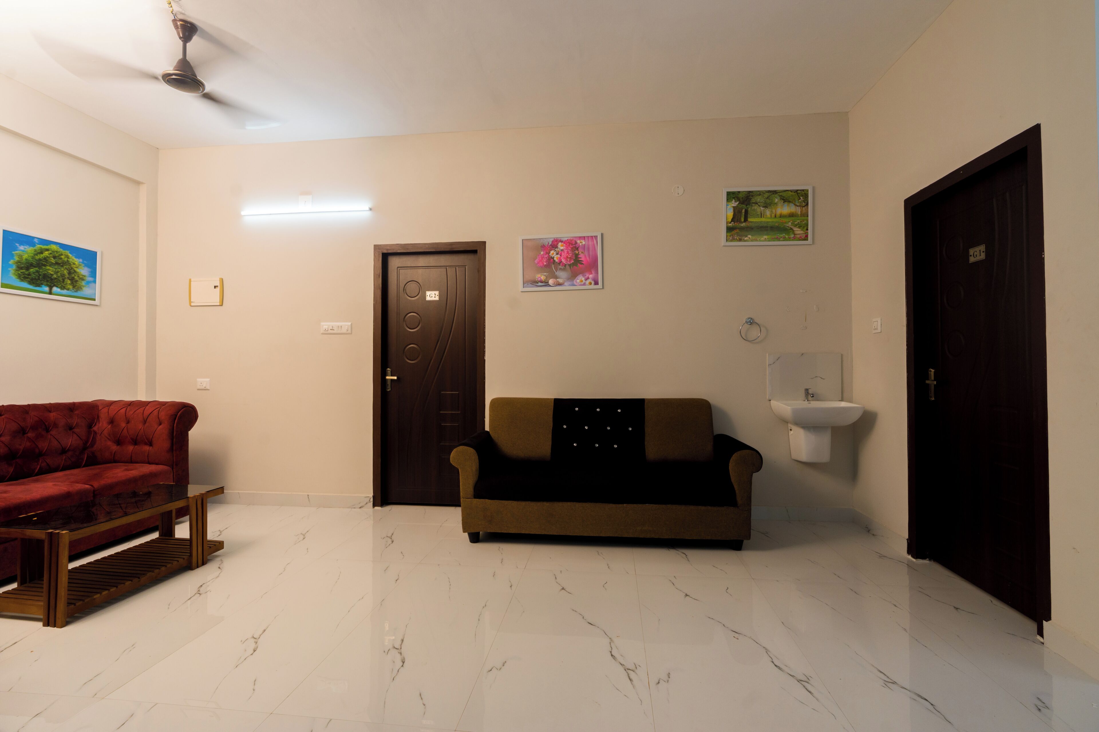Una habitación con un sofá rojo, un sofá marrón, una mesa de centro de madera, un ventilador de techo, una puerta y un lavamanos fijado a la pared.