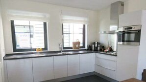 A modern kitchen with white cabinets, a built-in oven, a coffee maker, and a window with blinds. - Holiday Home in Uitdam With Wi-fi (Uitdam)