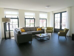Ein modernes Wohnzimmer mit einer Couch, Sesseln, einem Couchtisch und großen Fenstern.
