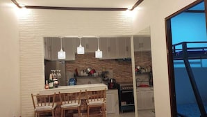 A compact kitchen with white cabinets, a brick backsplash, hanging pendant lights, and a dining area with wooden chairs.