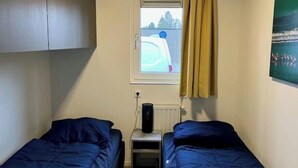 Ein kleines Zimmer mit zwei Einzelbetten, einem Fenster mit Blick auf einen Zaun, einem Heizkörper und einer Steckdose.