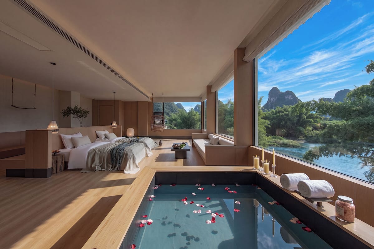 A modern spa room with a large window, a pool with rose petals, and a view of a river and mountains.