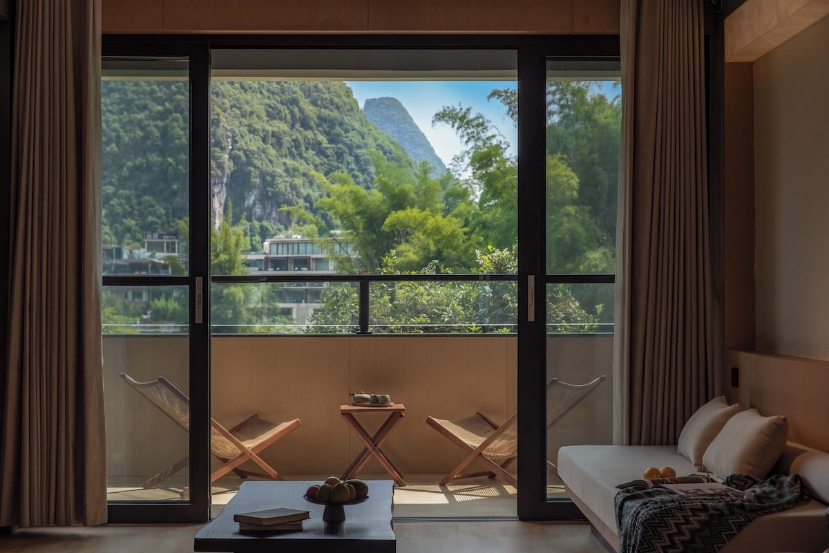 A room with a balcony, a sofa, a table with a vase, and a view of a mountain and trees.