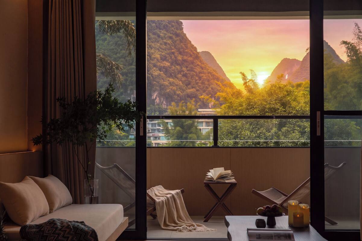 A room with a bed, a table with books and candles, and a view of mountains through large windows.