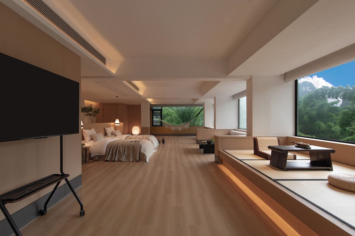 A modern hotel room with a large bed, seating area, TV, and a view of greenery.