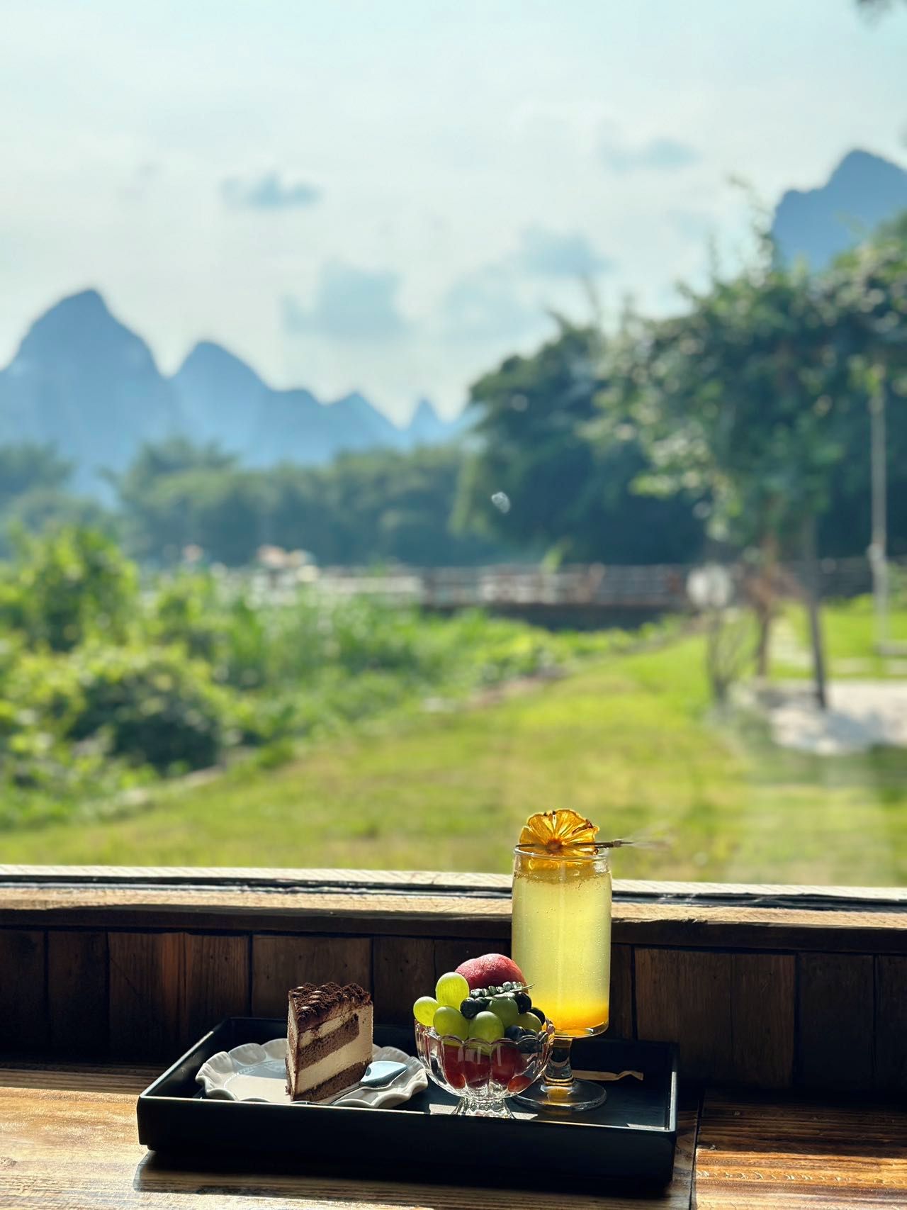 Un plateau de desserts avec une part de gâteau et un bol de fruits sur une table en bois, et un verre de boisson glacée au premier plan.