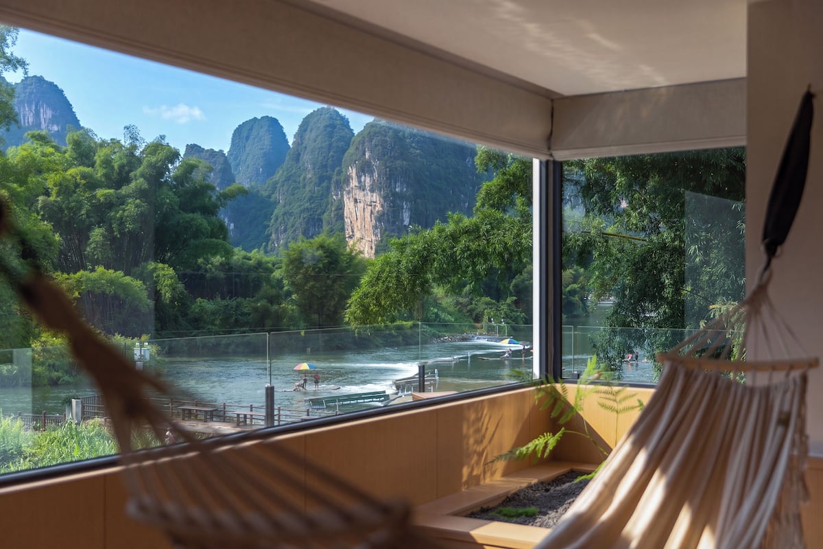A room with a large window overlooking a river and mountains. There is a hammock and a wooden bench.