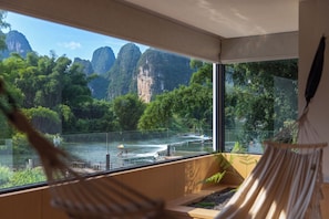 A room with a large window overlooking a river and mountains. There is a hammock and a wooden bench. - Qixi Meidi Yangshuo's Living Water (Guilin)
