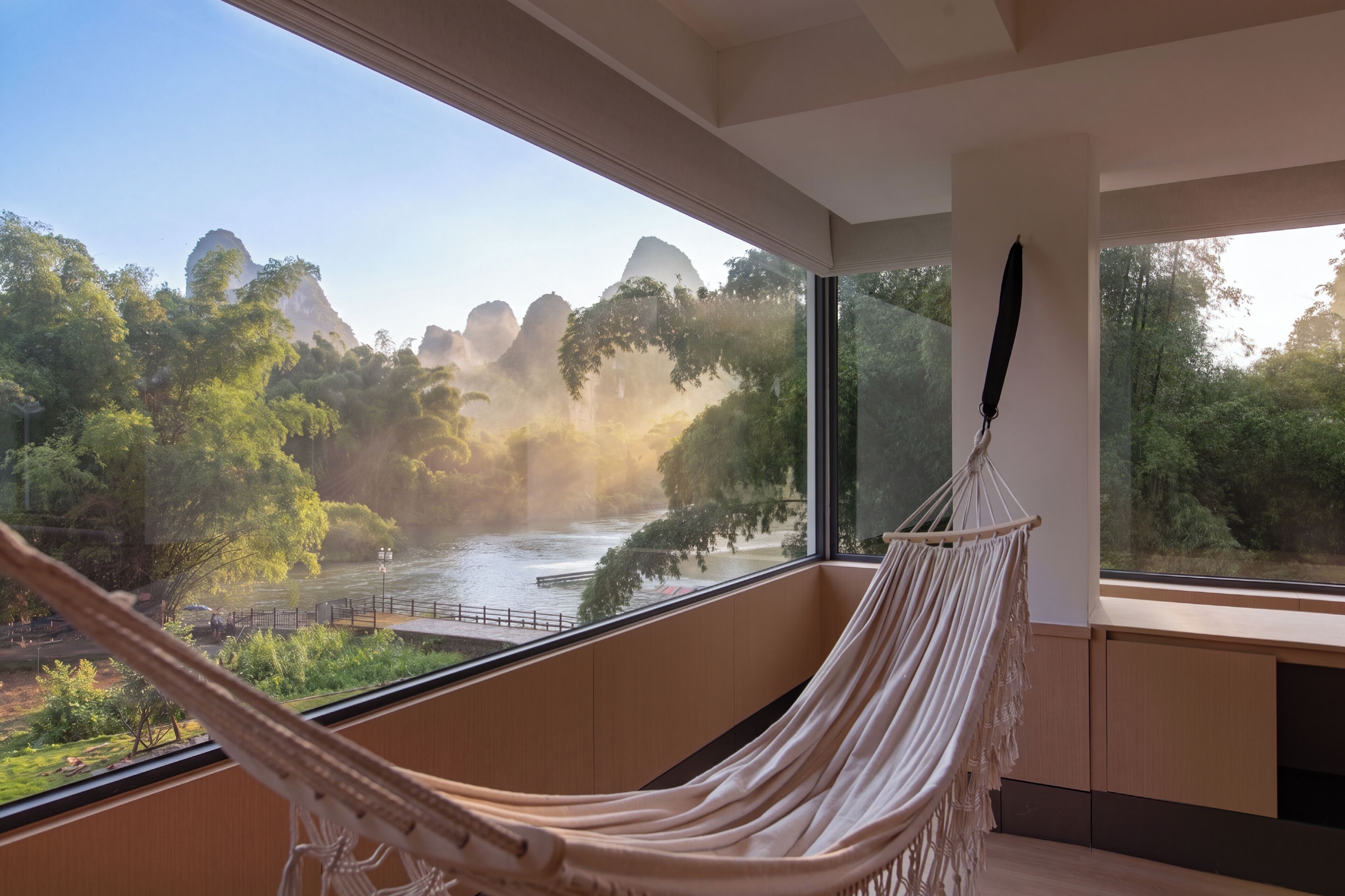 A hammock in a room with large windows overlooking a river and forested landscape.
