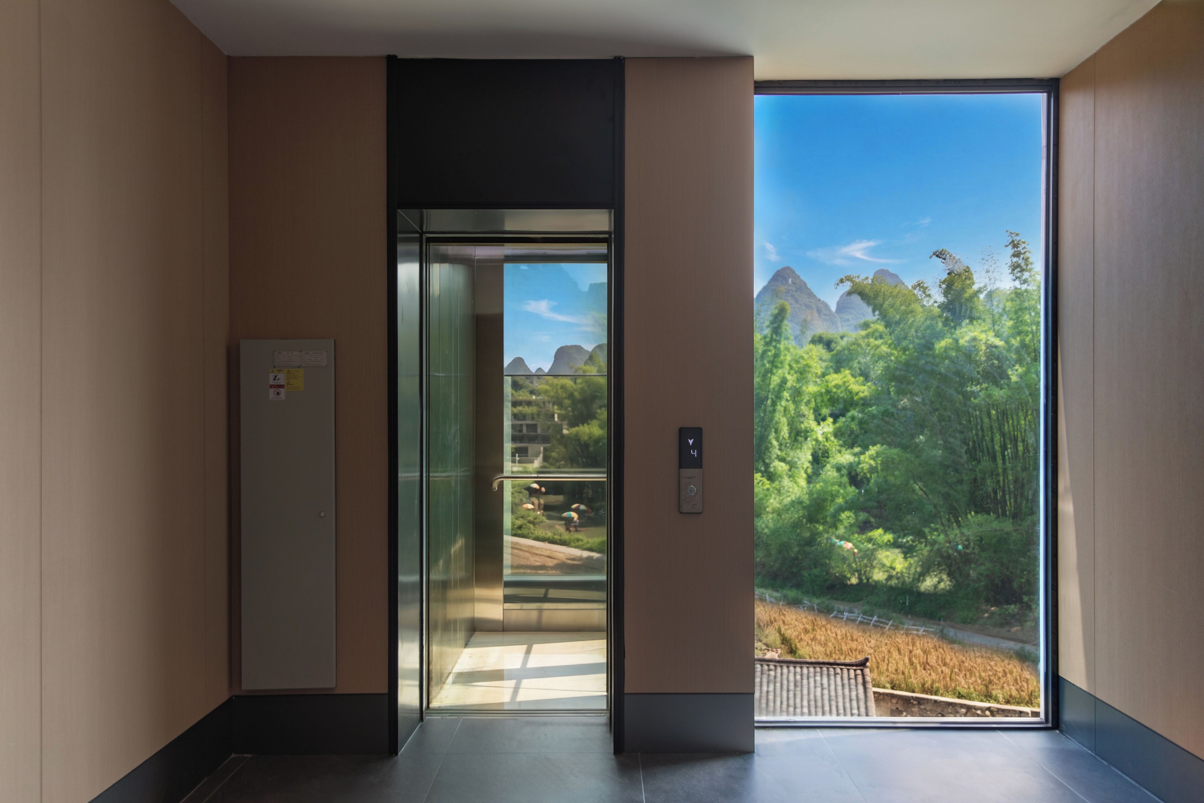 A modern elevator with a glass door, a control panel, and a view of a lush landscape with trees and mountains.