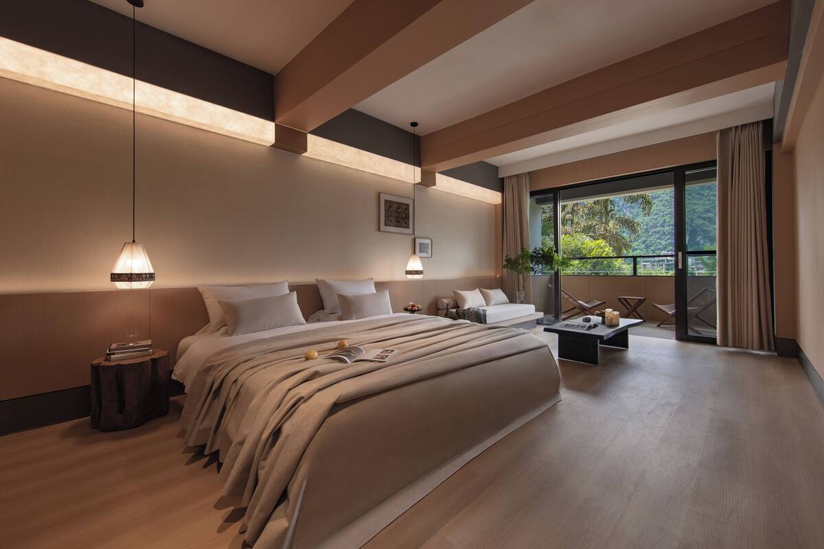 A modern bedroom with a large bed, bedside tables, a nightstand, a desk, and a window with a view of greenery.