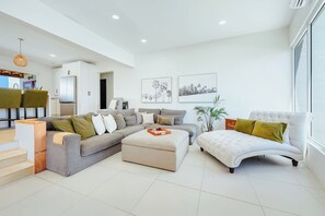 A modern living room with a grey sectional sofa, a white armchair, and a coffee table. There is a kitchen area visible in the background.