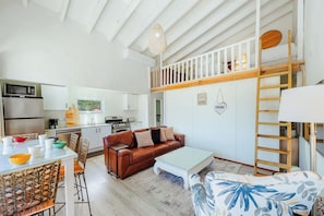 Un espacio habitable compacto con kitchenette, un sofá de cuero marrón, una mesa de centro de vidrio y una pared blanca con decoración en forma de corazón.
