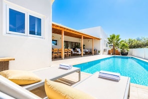 Un área moderna junto a la piscina con sillones de descanso, un espacio de estar cubierto y una piscina de aguas cristalinas.