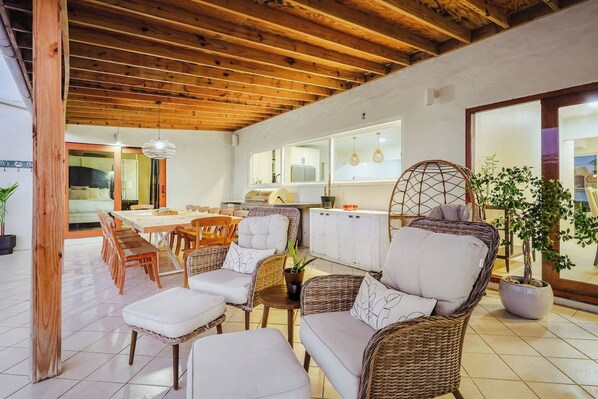 A spacious living area with a high ceiling and wooden beams, furnished with wicker sofas and chairs, a dining table, and a kitchen in the background.