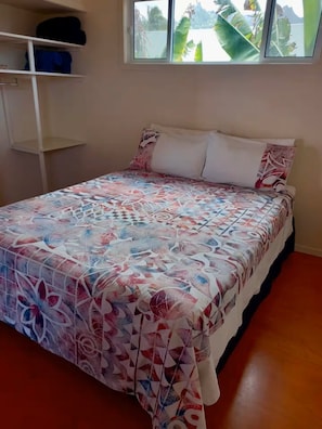A bed with a patterned bedspread, a window with a view of greenery, and a white shelf unit. - Kuki Beach Bach (Matavera District)