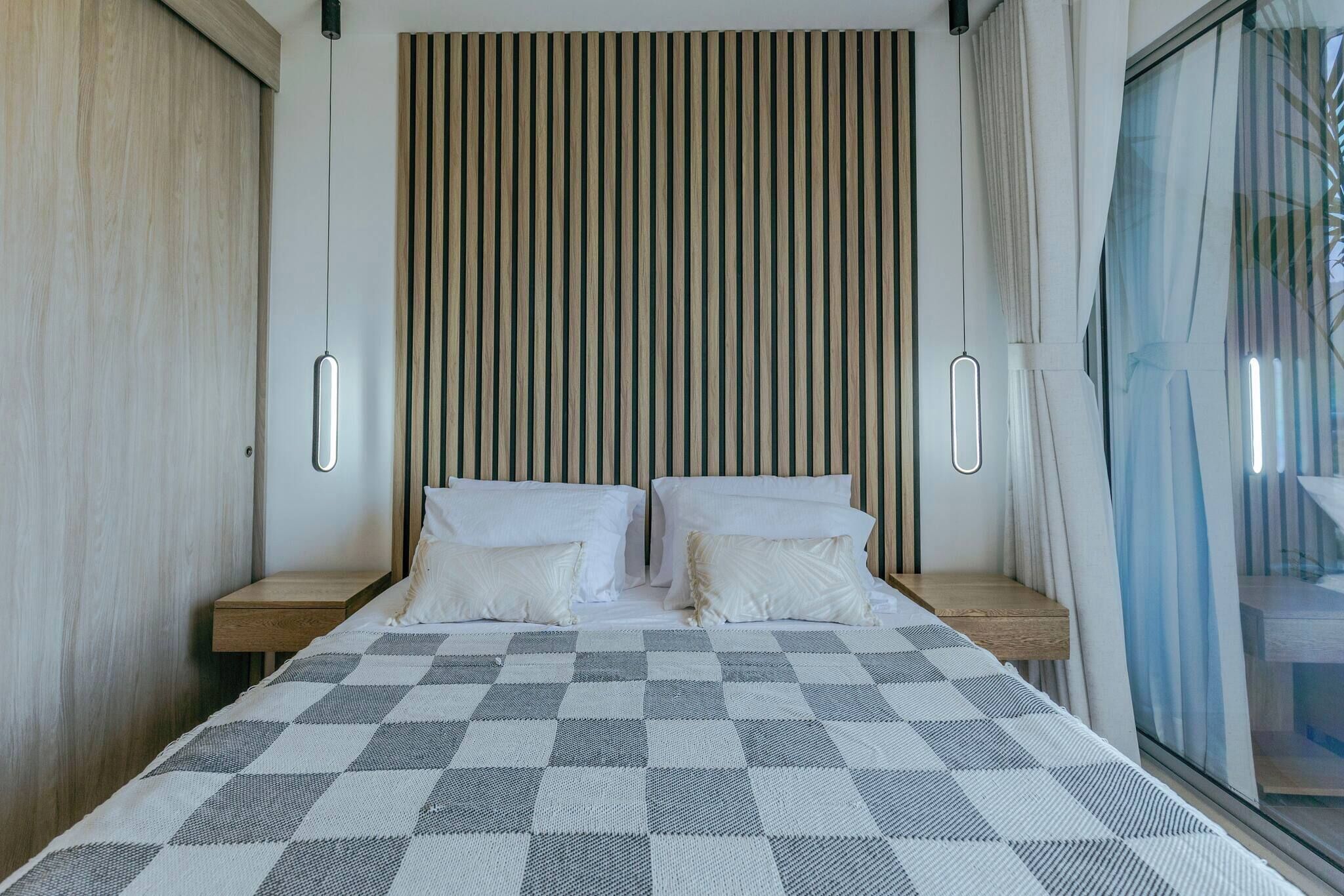A bedroom with a checkered bedspread, wooden slat headboard, bedside tables, and a view through a window.