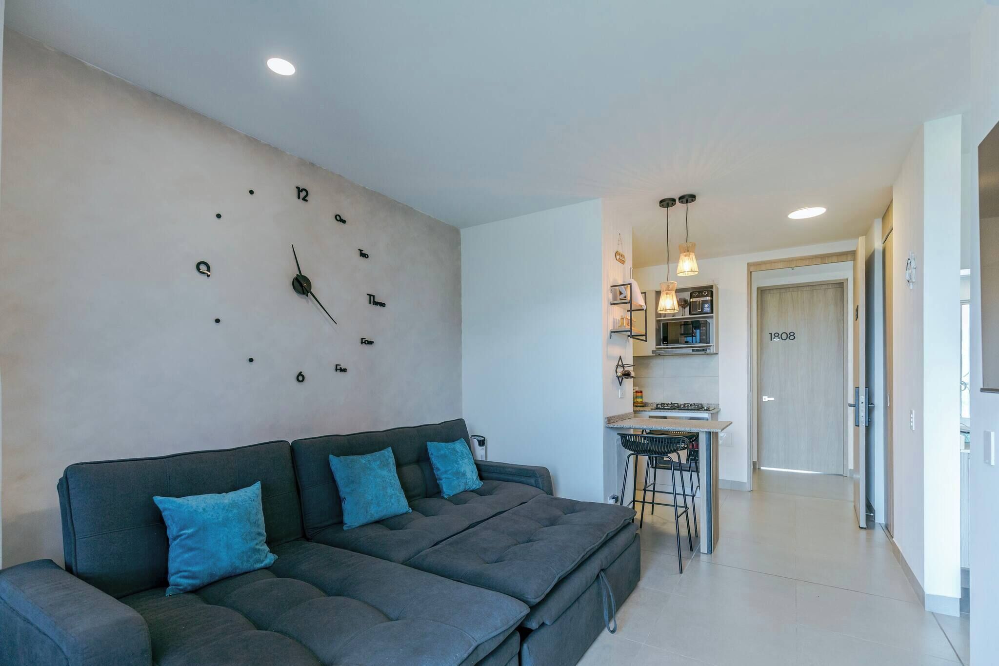 A modern living room with a sofa, a wall clock, and a kitchen area in the background.