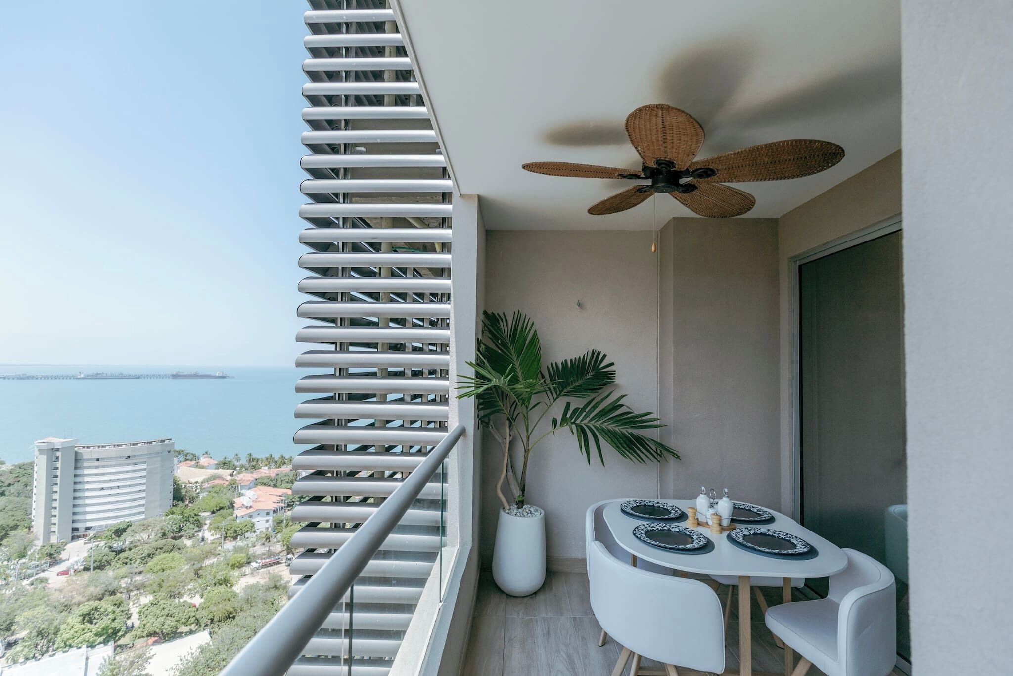 A balcony with a view of the sea, a ceiling fan, a potted plant, and a dining table with plates.