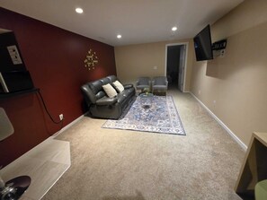 Een woonkamer met een leren bank, twee fauteuils, een kamerkleed met patroon en een flatscreen televisie aan de muur.