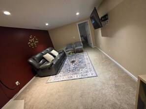 Een woonkamer met een leren bank, een kamerbreed tapijt, een flatscreen televisie aan de muur en een doorgang naar een andere kamer.