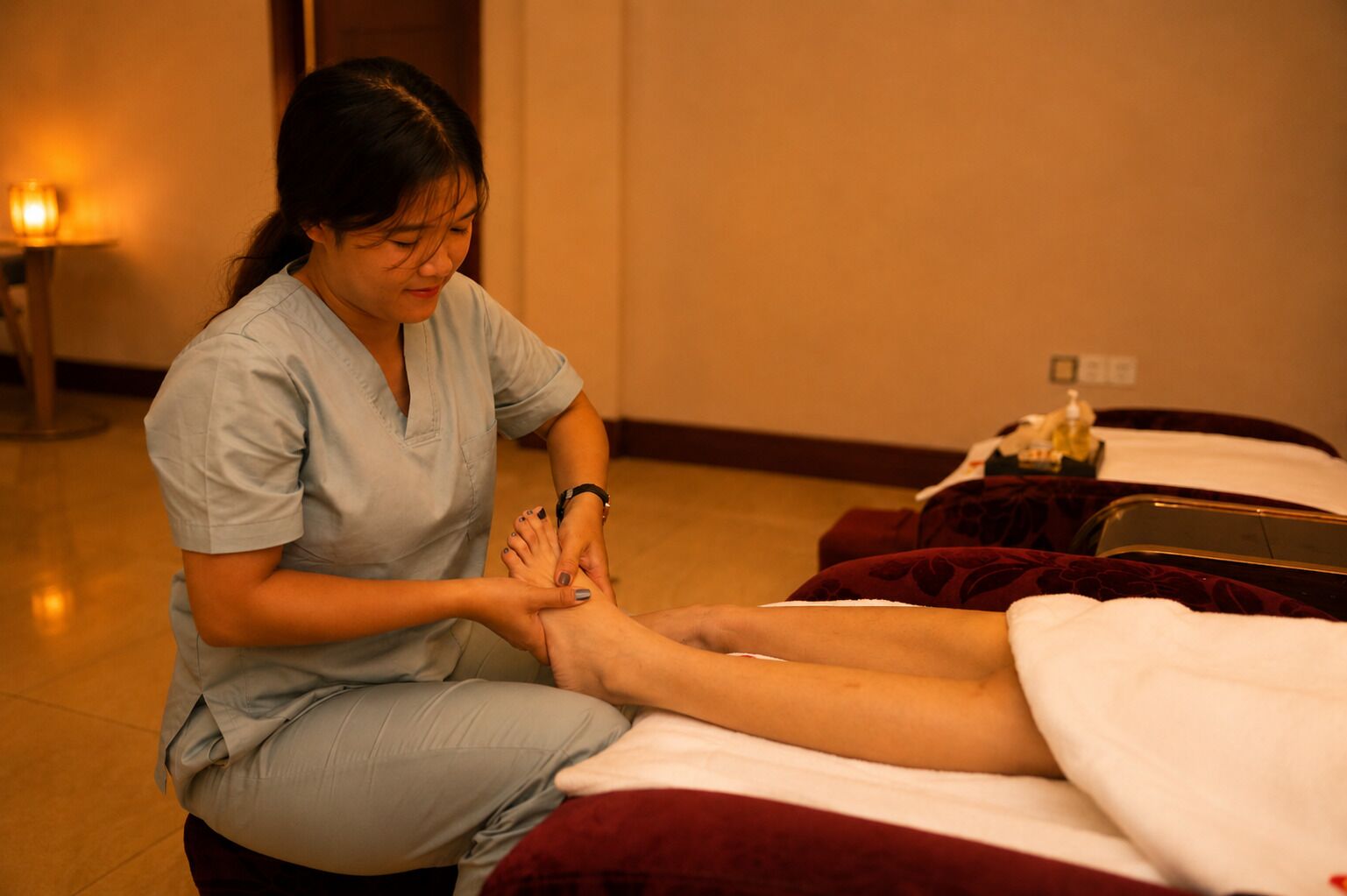 Une personne reçoit un massage des pieds dans une pièce où se trouvent un lit, une petite table avec une bouteille et une lampe fixée au mur.