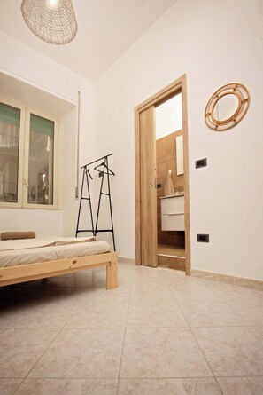 Una camera da letto con un letto, una finestra, una porta in legno, uno specchio e un appendi-asciugamani.