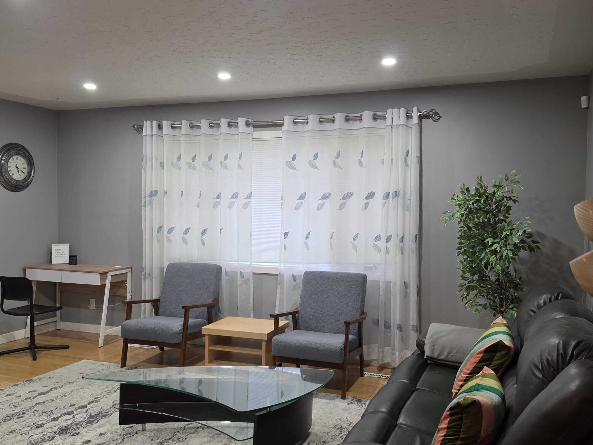 A living room with a glass coffee table, two grey armchairs, a wooden side table, a black leather sofa, a potted plant, and a white curtain with leaf patterns.