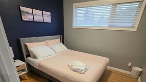 A bedroom with a bed, a nightstand, a window with blinds, and a wall-mounted artwork.