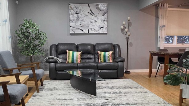 A living room with a black leather sofa, a glass coffee table, a grey armchair, a wooden side table, a potted plant, and a painting on the wall.