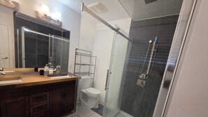 A bathroom with a glass-enclosed shower, a toilet, a sink with a mirror, and a shelf with toiletries.