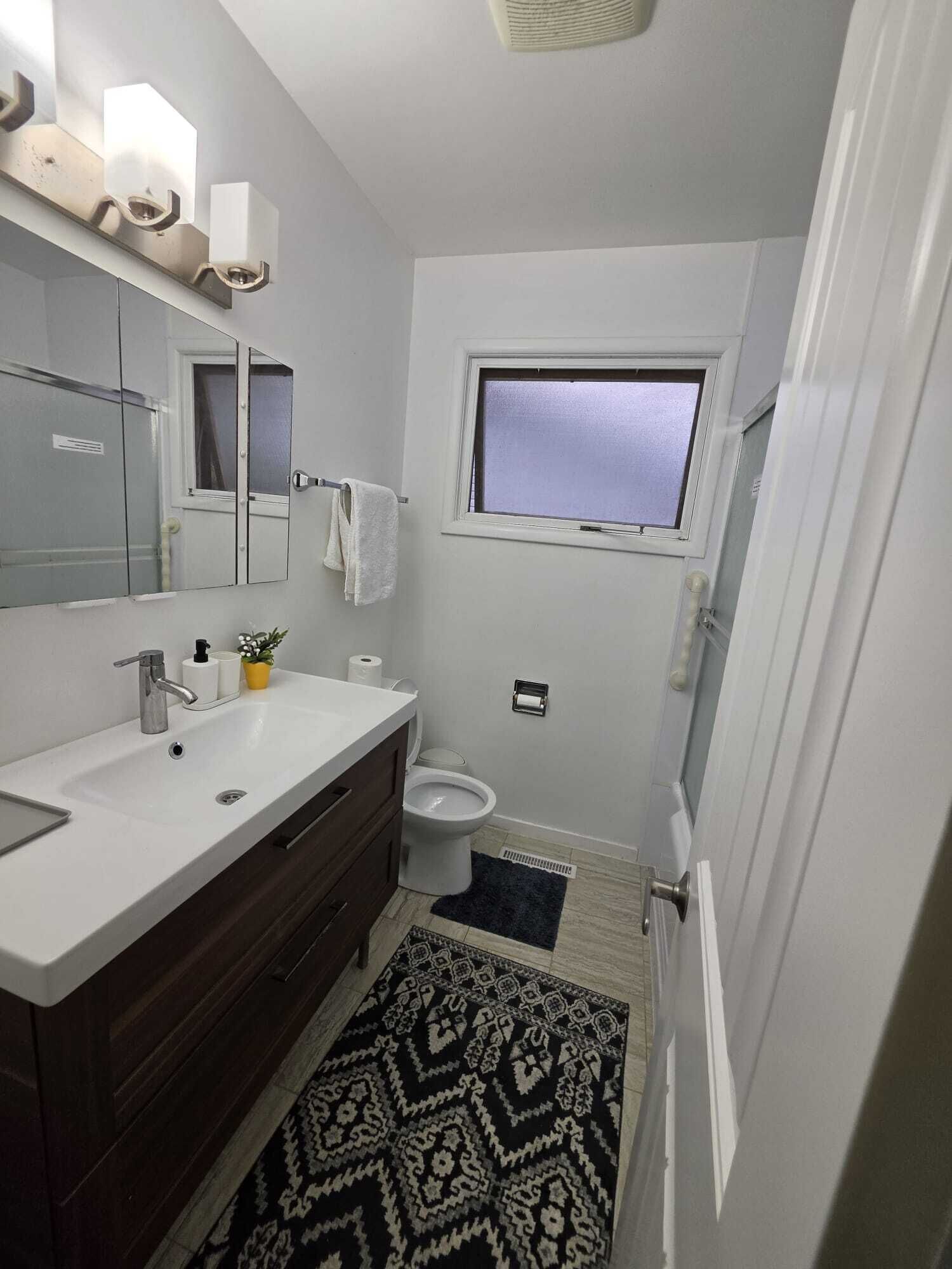 A compact bathroom with a white sink, dark wooden vanity, a mirror, a window, and a toilet.