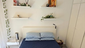 A bedroom with a bed, bedside table, lamp, and shelves with decorative items. - Fully furnished apartment, beaches, between Monaco and Menton (Roquebrune-Cap-Martin)