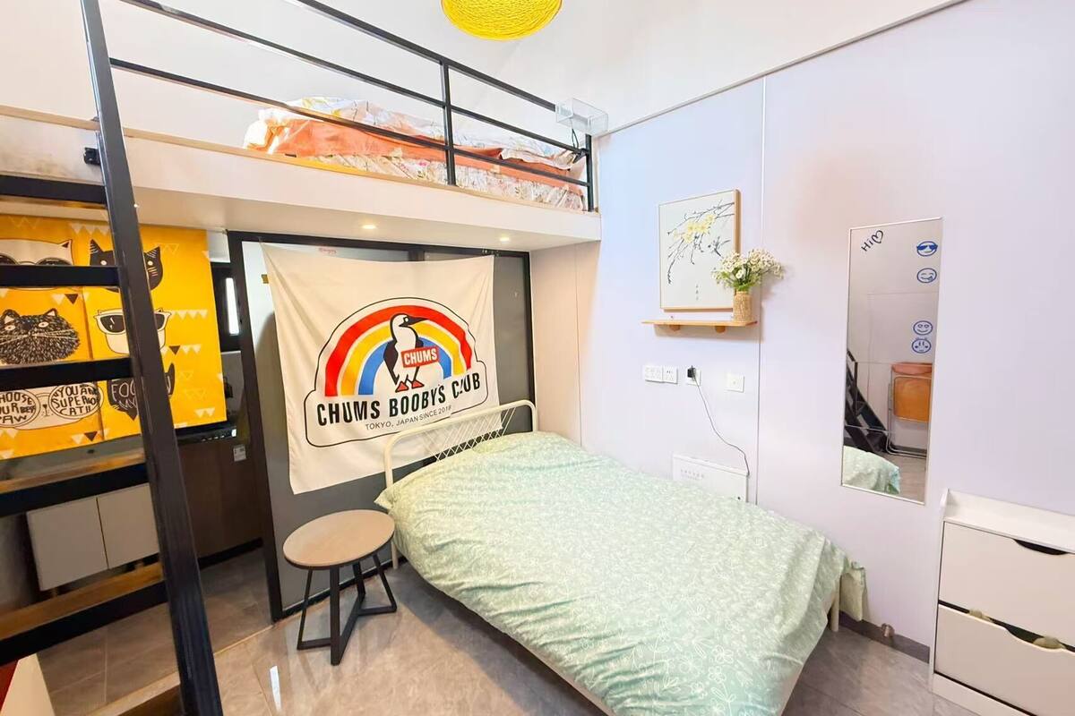 A bunk bed room with a bunk bed, a small table, a bed with a patterned bedspread, a wall-mounted shelf with a plant, and a wall with a framed picture and a sign.