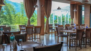 Un restaurante con mesas y sillas, amplias ventanas con cortinas y vistas a zonas verdes y montañas.