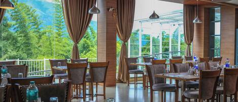 A restaurant with tables and chairs, large windows with curtains, and a view of greenery and mountains.