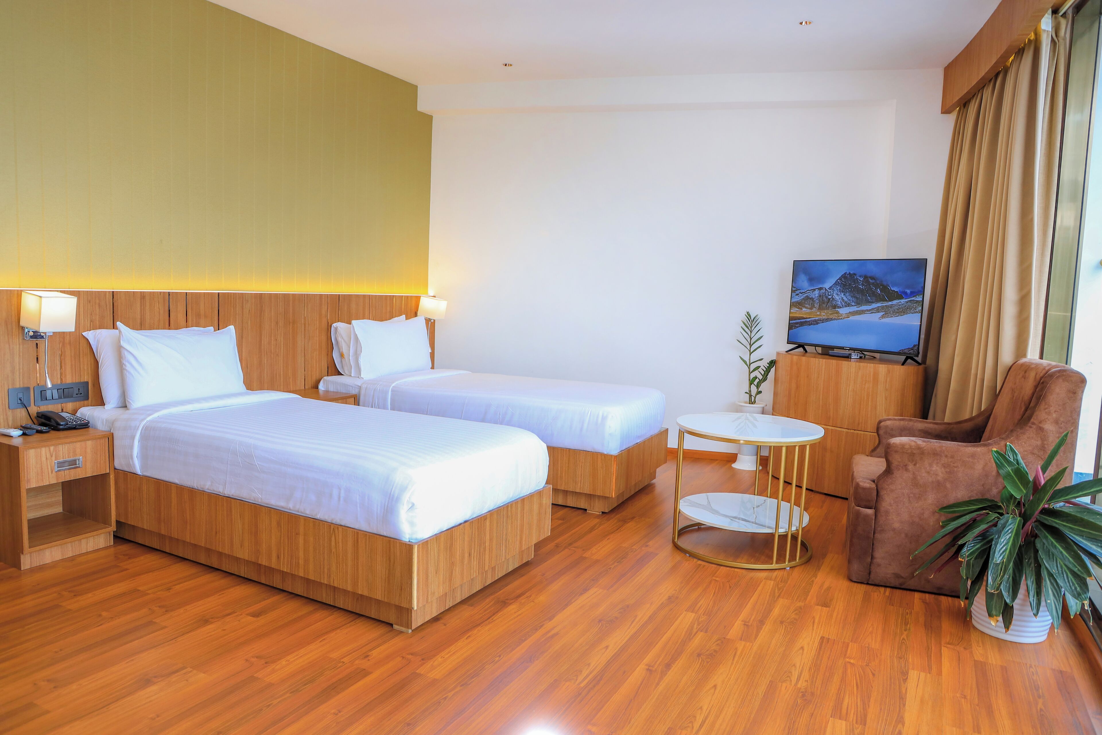 A hotel room with two beds, a TV, a chair, a small table, and a plant.