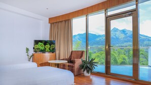 Habitación de hotel con cama, televisor, silla, mesita pequeña y un amplio ventanal con vista a la montaña.