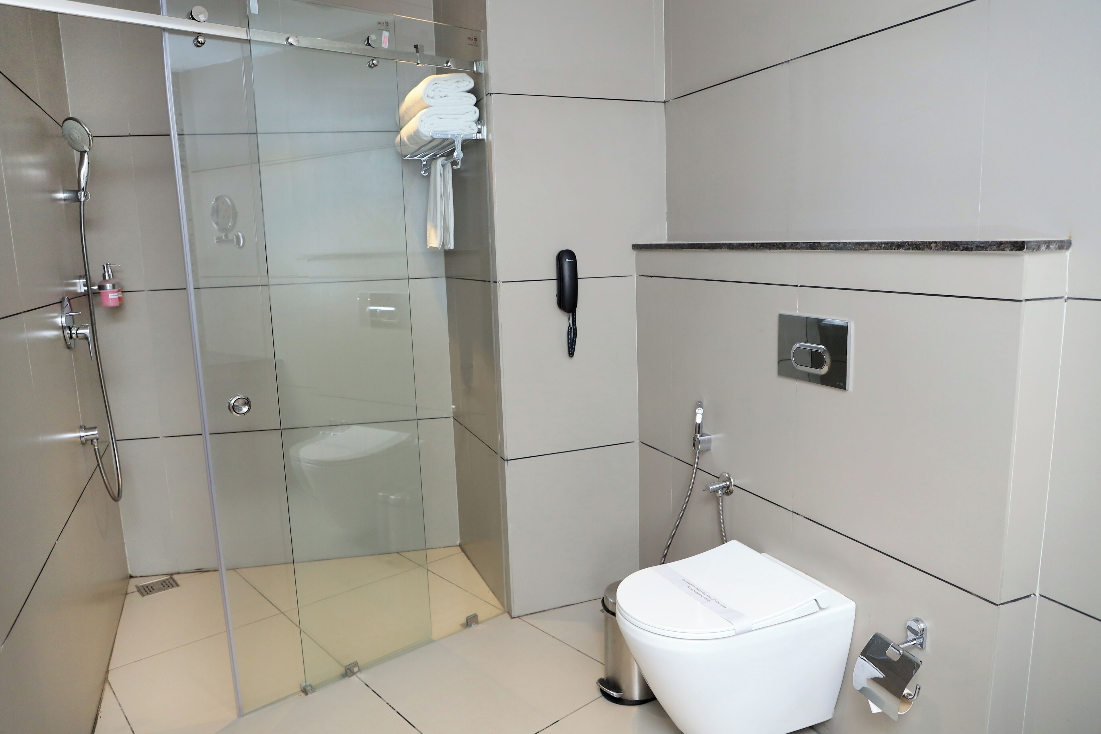 A modern bathroom with a glass shower enclosure, a white toilet, a trash bin, and a telephone mounted on the wall.