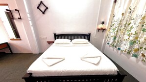 Una cama con colchón blanco y cabecera negra, una mesita de noche pequeña y un estante en la pared con una lámpara.