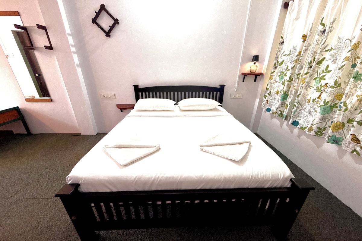 Una cama con colchón blanco y cabecera negra, una mesita de noche pequeña y un estante en la pared con una lámpara.