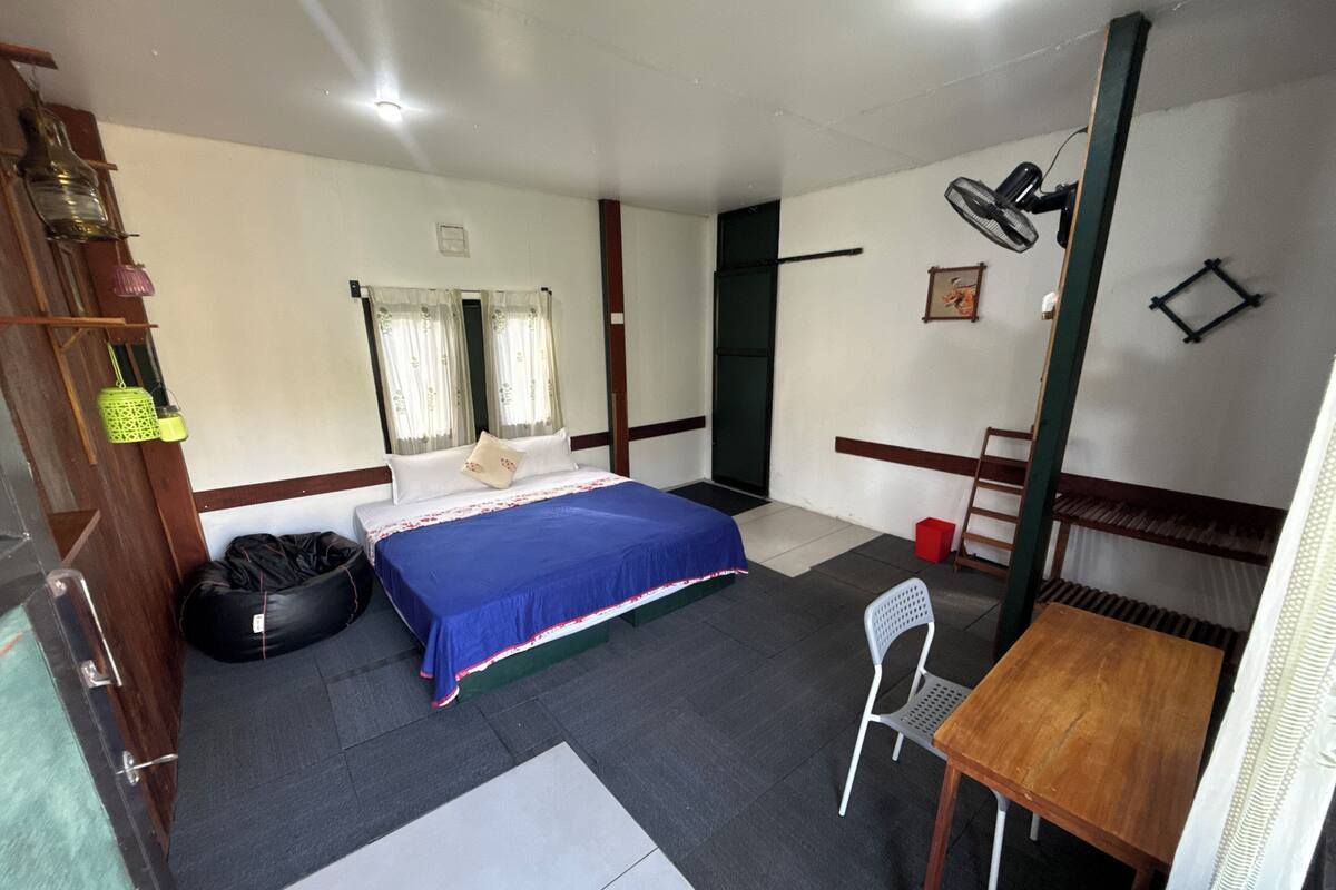 Una habitación con cama, mesa, silla, ventilador y escalera.