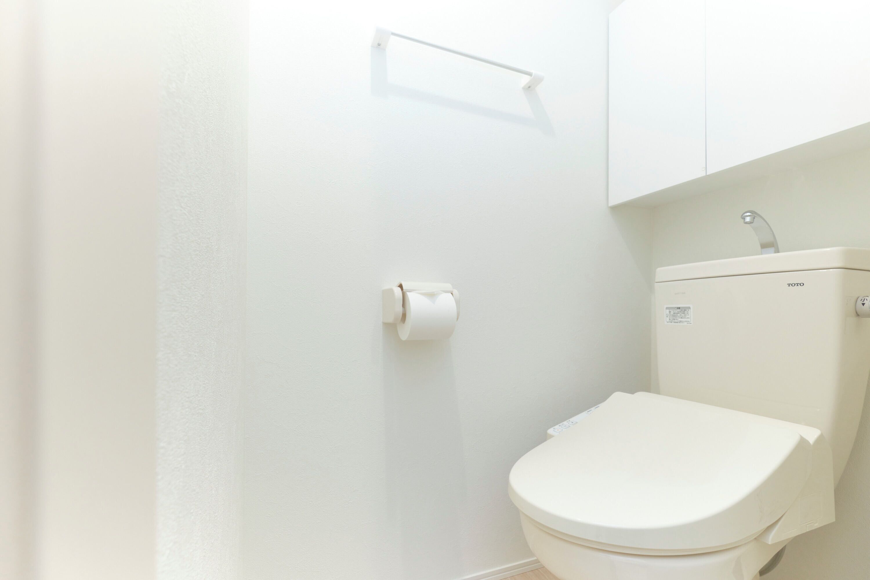 A white toilet with a TOTO brand label, a toilet paper holder on the wall, and a white cabinet above the toilet.