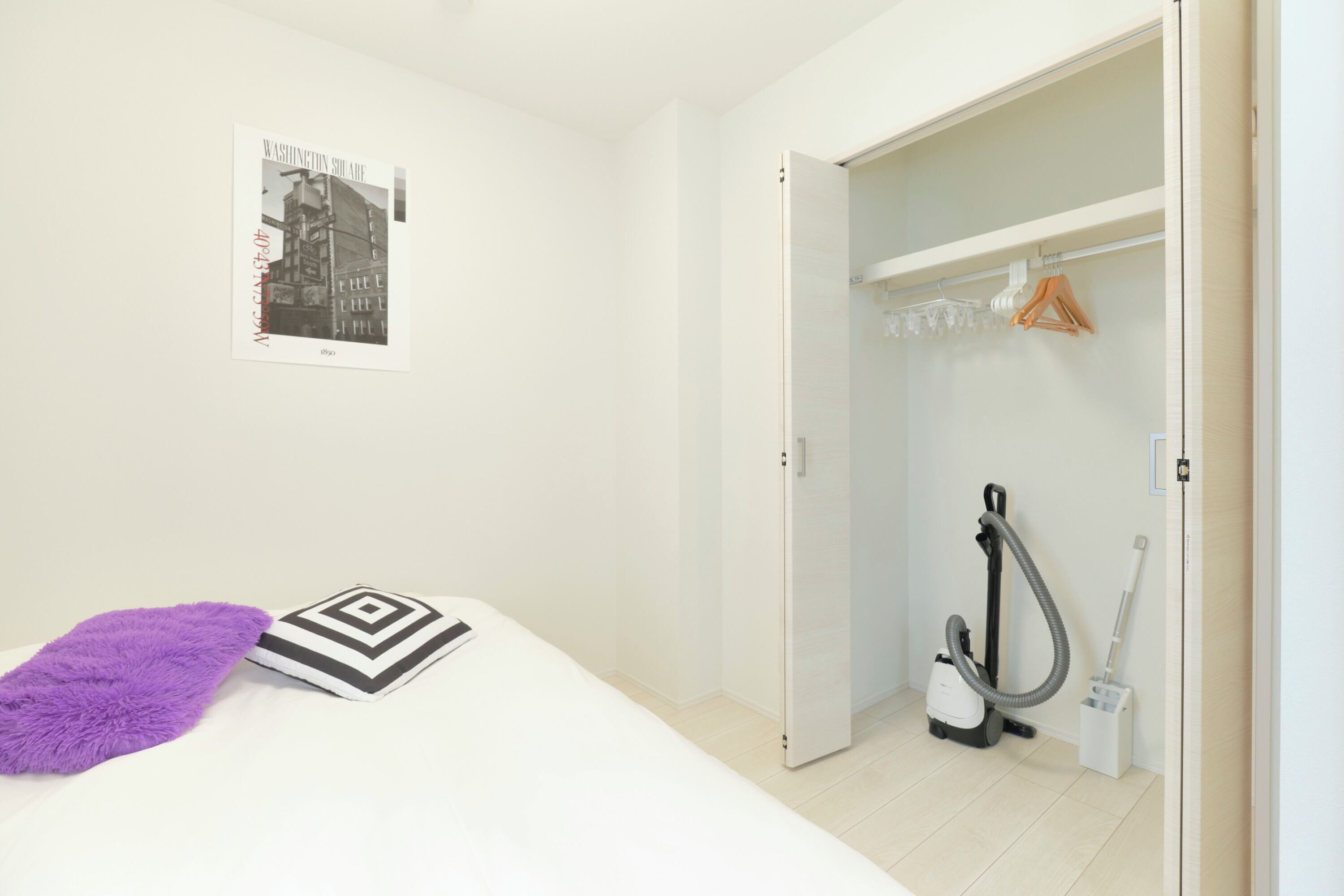 Een slaapkamer met een bed, een paarse plaid, een zwart-wit kussen, een stofzuiger en een inloopkast met hangende kleding.