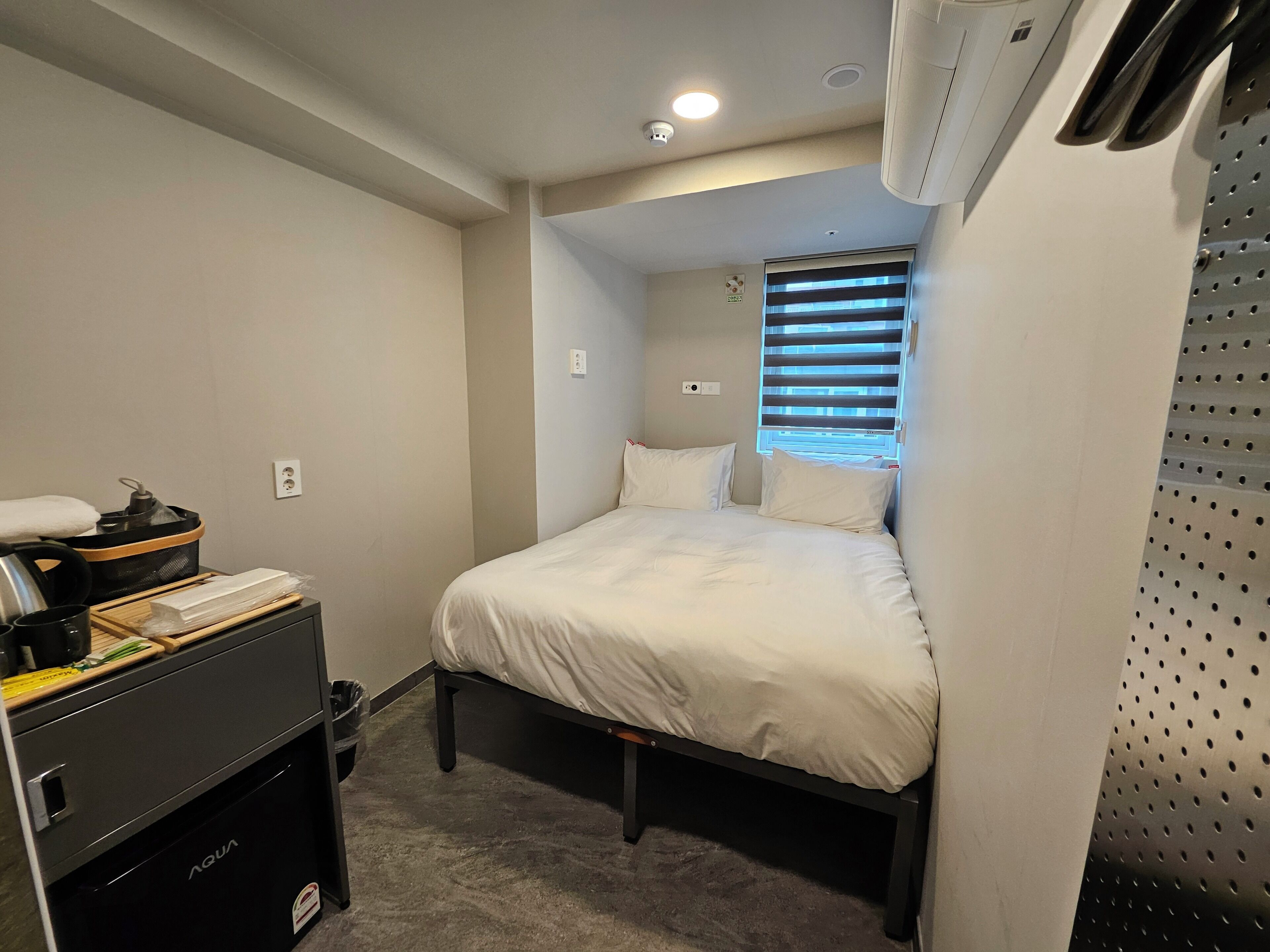 Ein kleines Hotelzimmer mit Bett, einem kleinen Tisch mit Wasserkocher, einem Mülleimer und einem Fenster mit Jalousien.