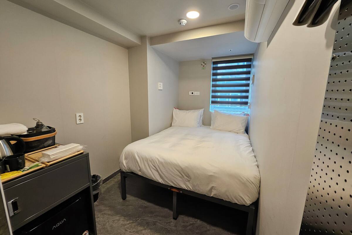 A compact hotel room with a bed, a small table with a kettle, a trash bin, and a window with blinds.