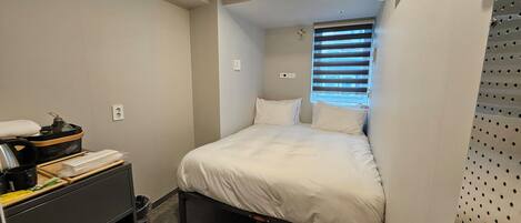 Habitación de hotel pequeña con cama, una mesita con hervidor, un cesto de basura y una ventana con persianas.